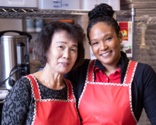 An Asian American and a Black woman working in the kitchen and smiling at the camera