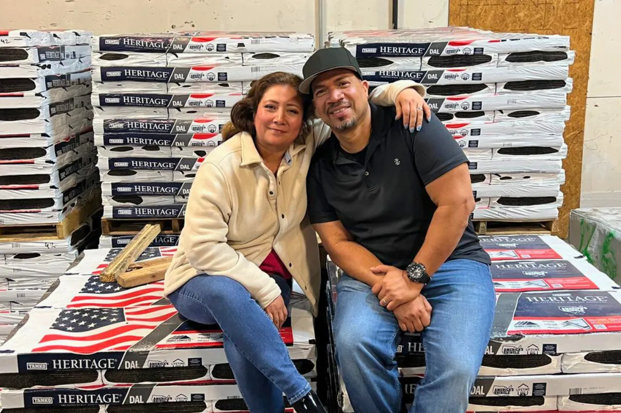 Esmeralda and Octavio Polanco joyfully sitting atop stacked roofing supplies in their new warehouse, sharing a moment of laughter and enjoyment together amidst their small business success.