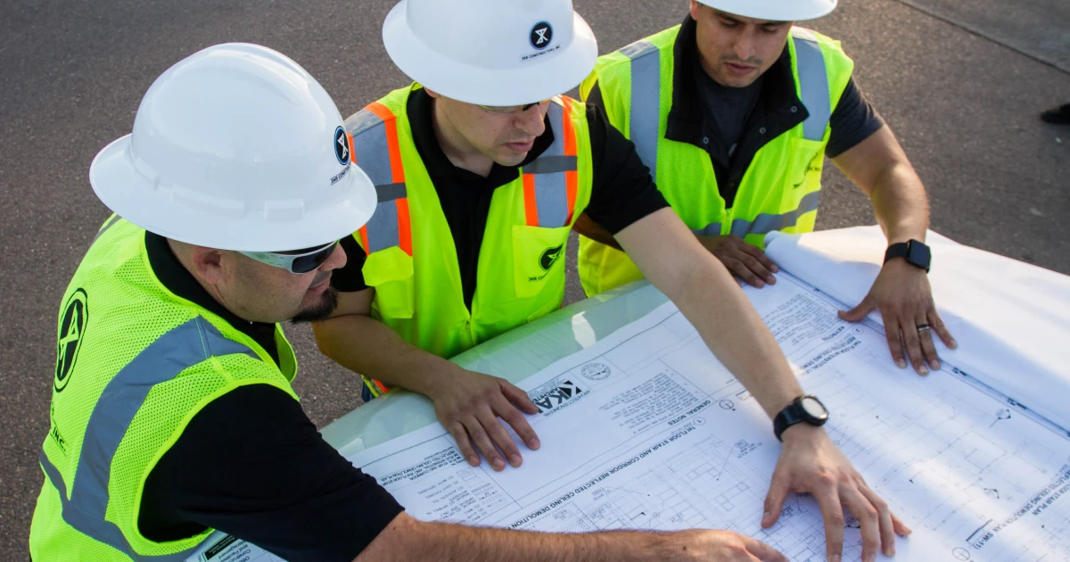 Three men in hard hats examining blueprints at a construction site, discussing project details and plans.